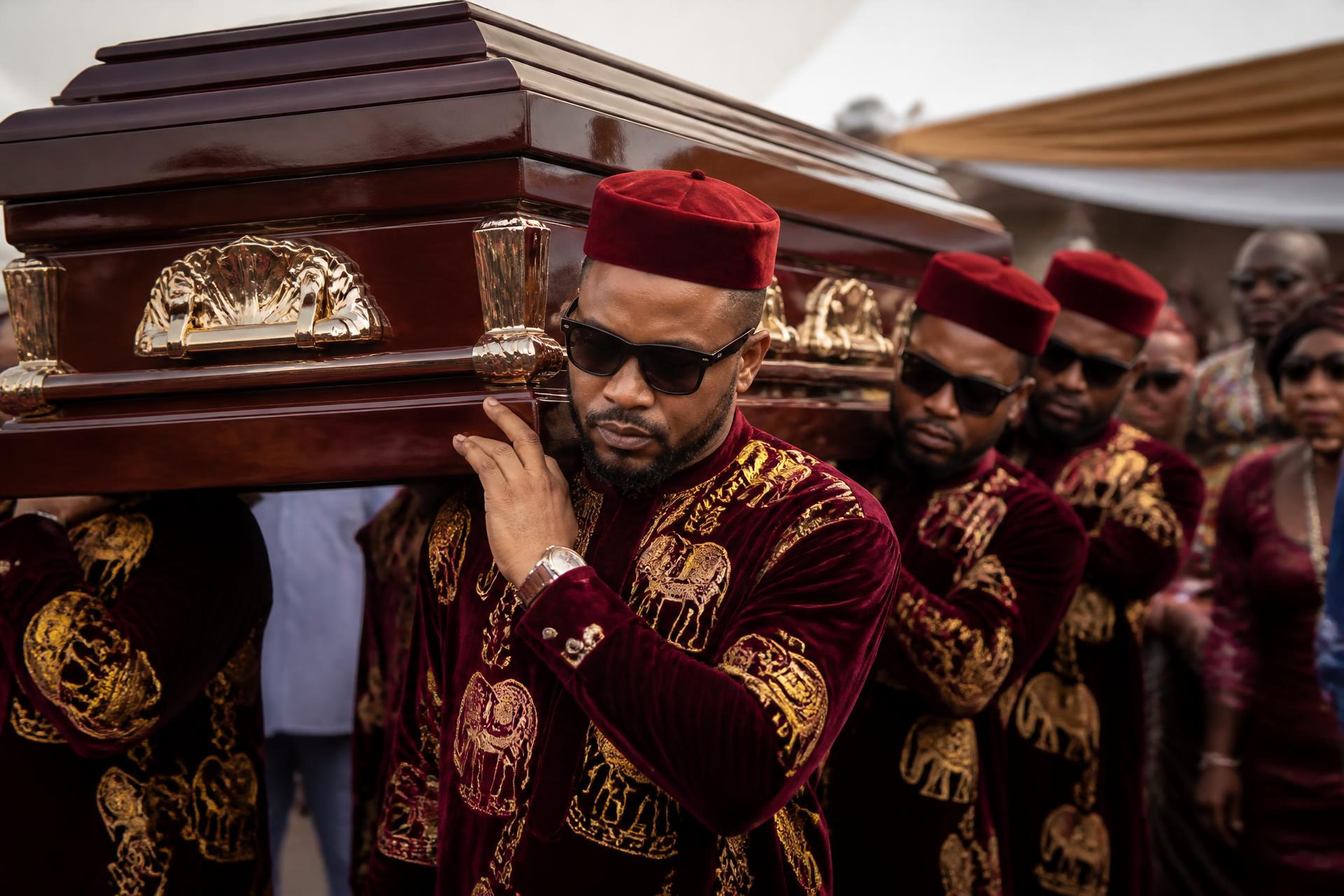 Professional Igbo funeral undertakers in burgundy and gold traditional attire carrying an elegant casket during a dignified Nigerian funeral ceremony